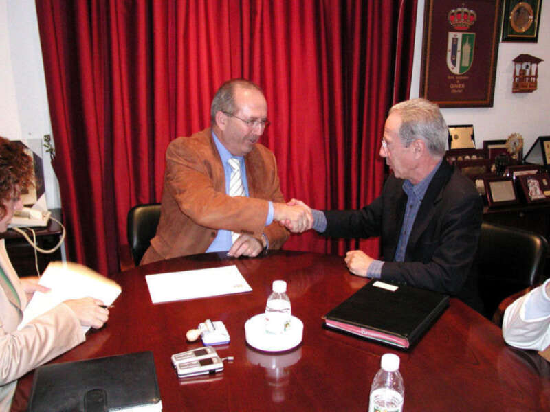 Dos hombres se saludan en una reunión formal, sentados frente a un escritorio con documentos y un teléfono móvil. La imagen muestra una atmósfera de negociación o acuerdo entre los participantes.