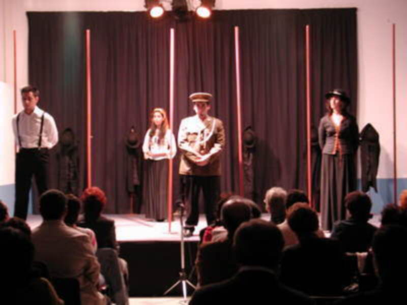 Una escena teatral con actores en trajes formales, un hombre y una mujer frente a la pantalla. La audiencia observa desde el fondo.