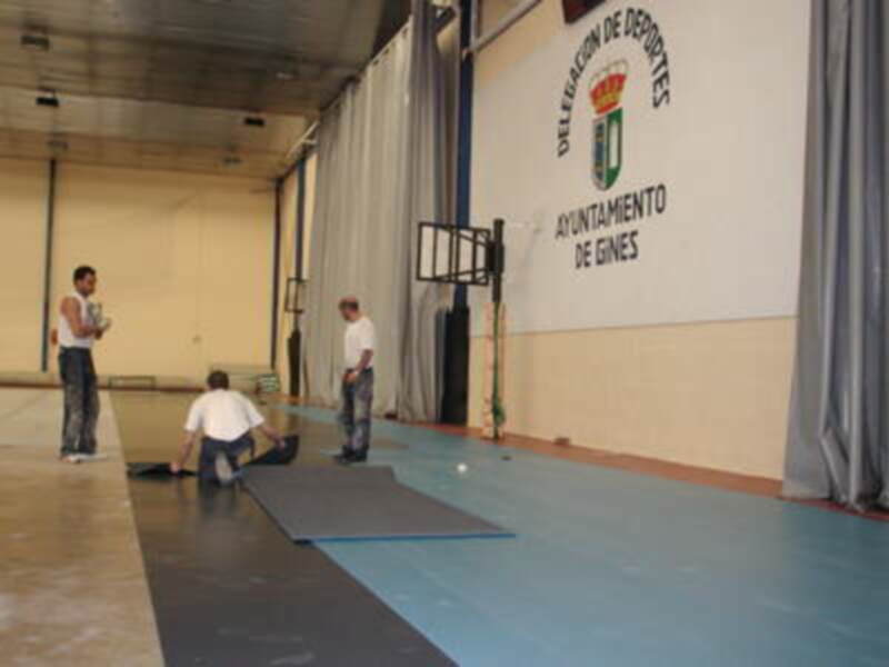 En un gimnasio de la Delegación de Deportes del Ayuntamiento de Gines, se observan dos hombres trabajando en el suelo. La pared muestra la bandera del municipio y un logo de la institución.