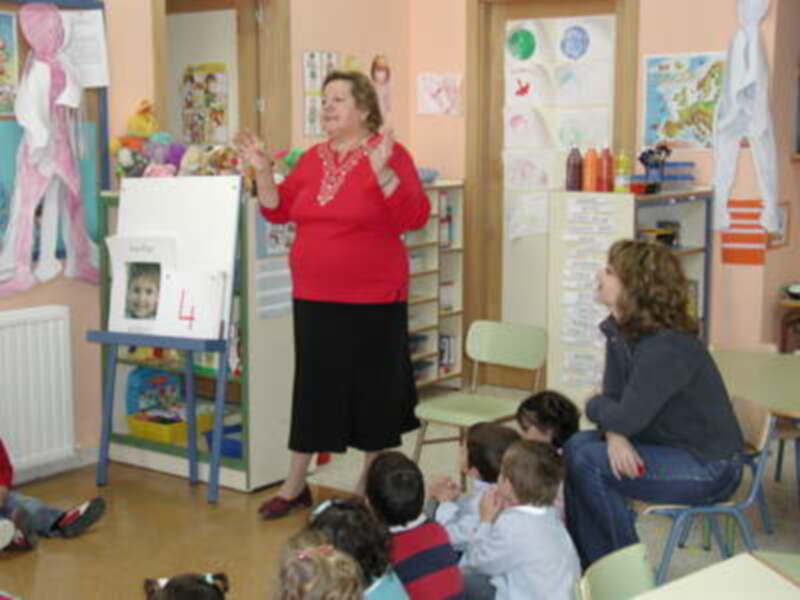 Una maestra en un aula con niños escuchando. En el fondo, se pueden ver estanterías con libros y cuadros decorativos.