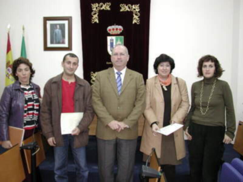 Una persona sostiene un diploma mientras otros tres asistentes observan. En el fondo, hay una bandera y un retrato de un hombre enmarcado.