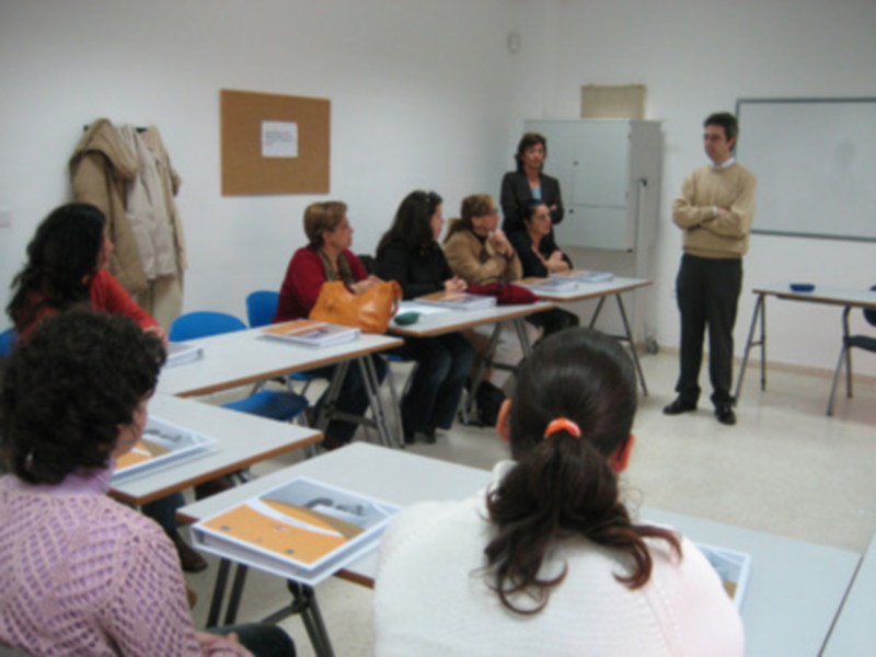 Una clase en un aula con estudiantes sentados en mesas, una persona enseñando y otros observando. La imagen muestra un ambiente educativo con libros y material de aprendizaje en la mesa del frente.
