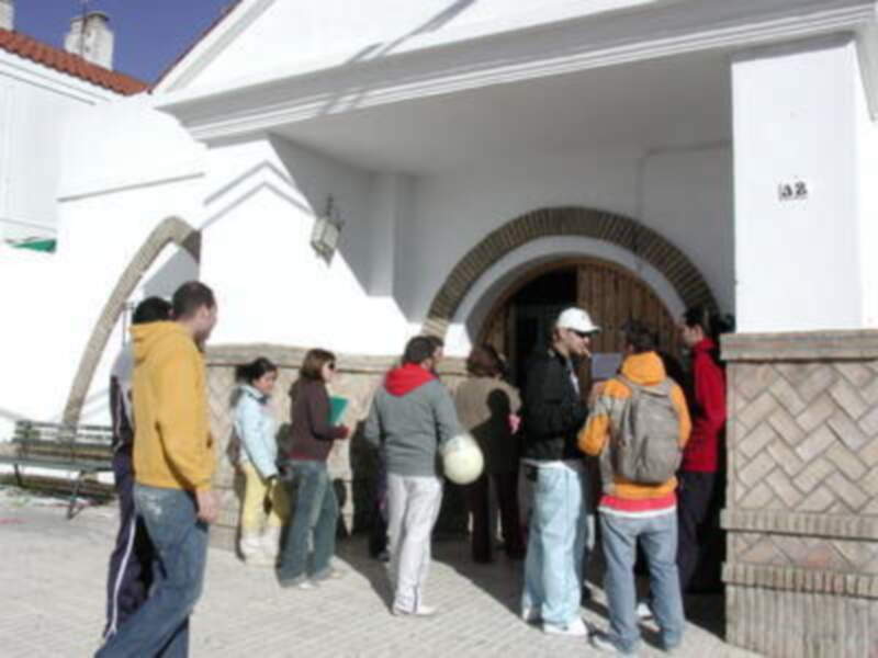 Gente esperando en línea frente a una estructura blanca con arcos y ventanas.