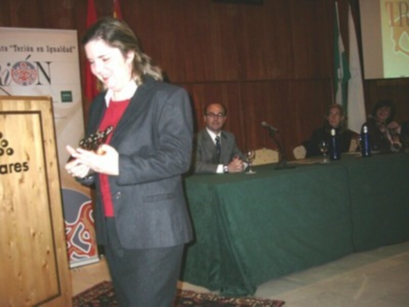 Una mujer en traje negro y corbata roja está hablando en un evento formal, con un hombre detrás de ella en traje y corbata, y otros asistentes en el fondo.