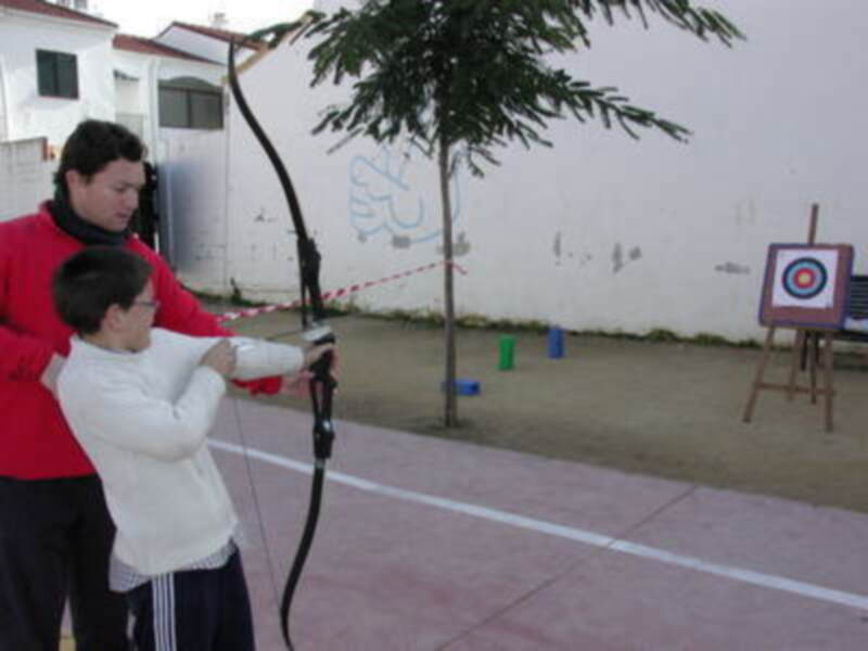 Una persona mayor y un niño están aprendiendo a arco y flecha en una zona de práctica al aire libre. El niño sostiene un arco mientras la persona mayor lo ayuda, y en el fondo se ve una caja de flechas con un objetivo.