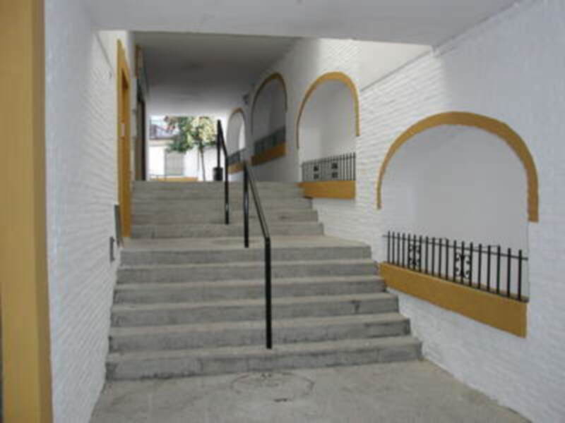 Escaleras con barandilla negra, paredes blancas y detalles amarillos. Arcos decorativos en las escaleras. Piso de hormigón claro. Entrada a un edificio con ventanas y plantas exteriores visibles.