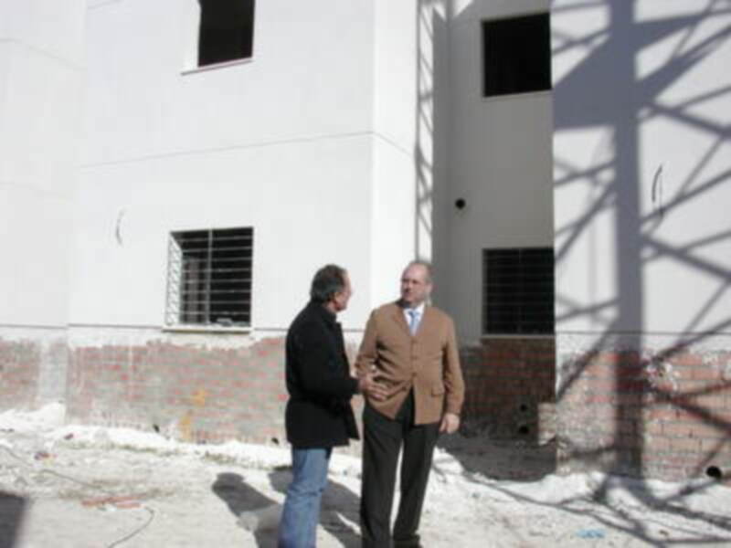 Dos personas conversando frente a una construcción en construcción, con una fachada blanca y una base de ladrillos rojos. El entorno parece ser un área urbana en construcción o rehabilitación.