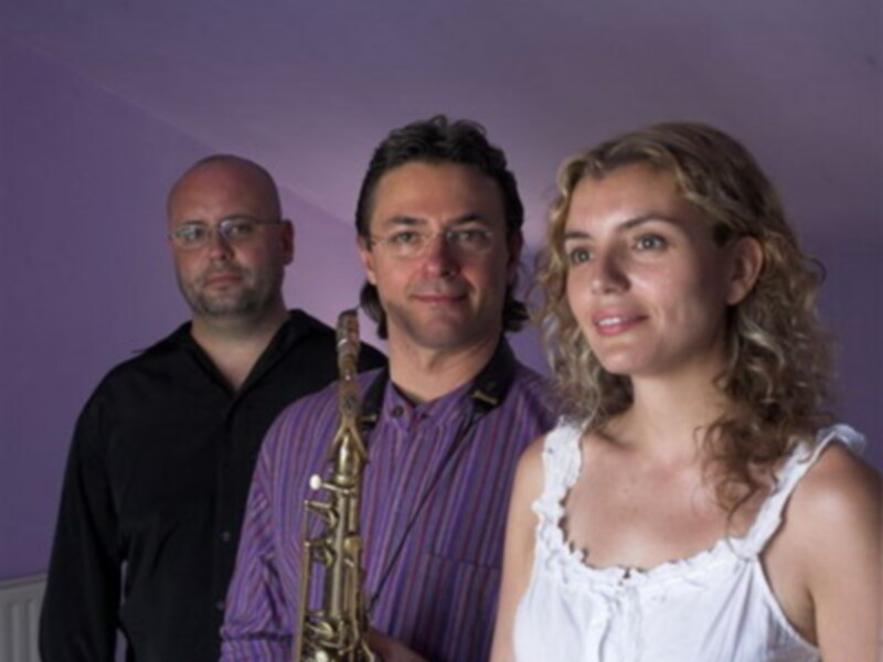 Tres personas posan frente a una pared púrpura, uno sostiene un saxofón.