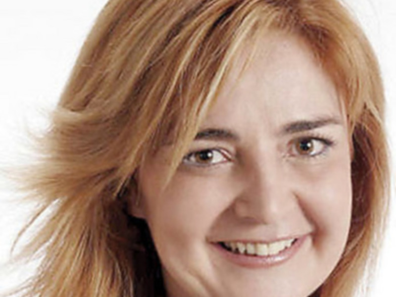 A una mujer con cabello largo y ondulado de color claro. Su expresión es amigable y sonríe con dientes visibles. La iluminación es suave, enfocándose en su rostro.