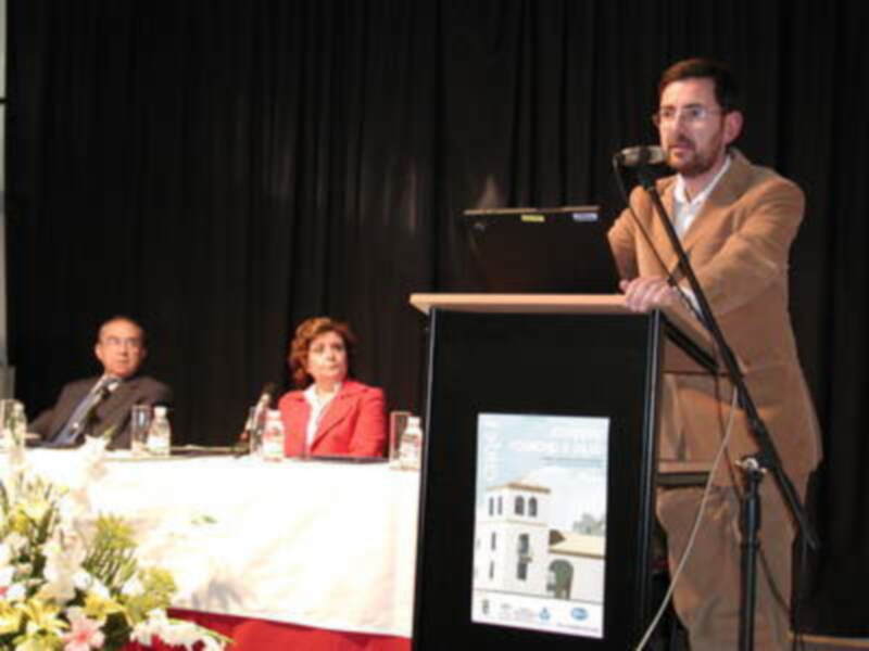 Un hombre habla en un podium, mientras una mujer y otro hombre escuchan. La imagen muestra una conferencia o evento formal con un fondo negro y un microfono en el podium.