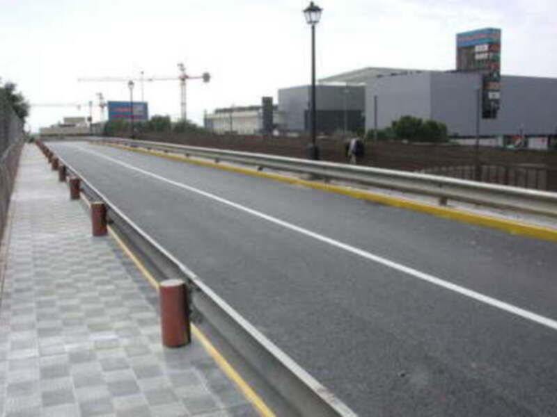 Carretera con carriles dobles, separados por una barrera de seguridad. A la izquierda hay un camino peatonal con adoquines, y a la derecha se observa una valla de seguridad. En el fondo hay edificios y un poste de luz.
