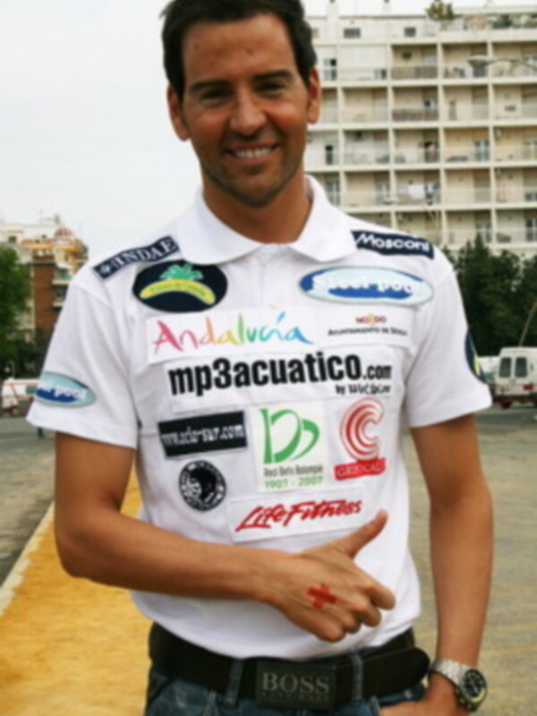 Un hombre sonríe mientras posa con un polo blanco lleno de logos y patrocinadores, incluyendo "Andalucía", "mp3acuatico.com" y "LifeFitness". Detalles como la correa de cuero del polo, el cinturón y el reloj de pulsera se destacan.