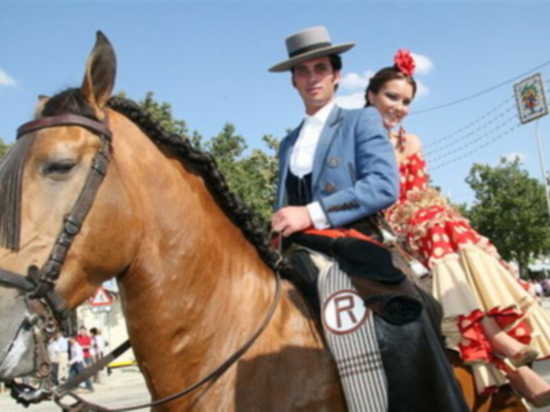 Elegante pareja en trajes típicos andaluces, montando a caballo en una celebración cultural.