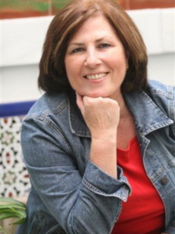 A una mujer con cabello marrón corto, sonriendo y apoyando la mano en la barbilla. Lleva una chaqueta vaquera azul y un top rojo. El fondo es de color beige con detalles en verde y marrón, posiblemente una pared o un mural.