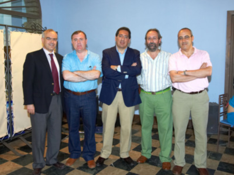 Cinco hombres de negocios posan juntos con brazos cruzados en una sala con suelo a cuadros y paredes azules.