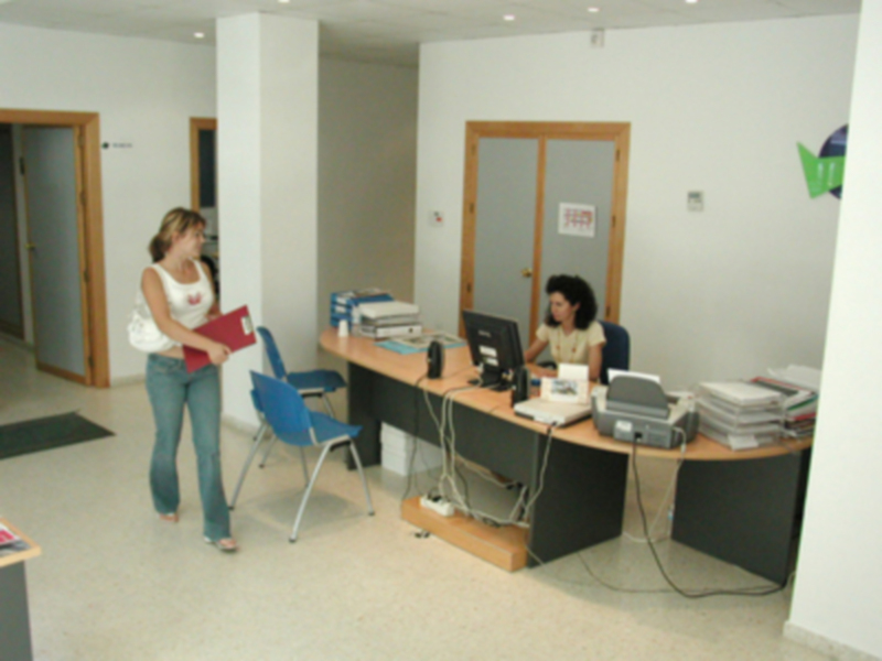 Una mujer camina con un bolso blanco y una carpeta roja, mientras otra trabajadora se sienta en una oficina con un escritorio lleno de documentos y equipos. La oficina tiene puertas correderas, una impresora y varios escritorios.