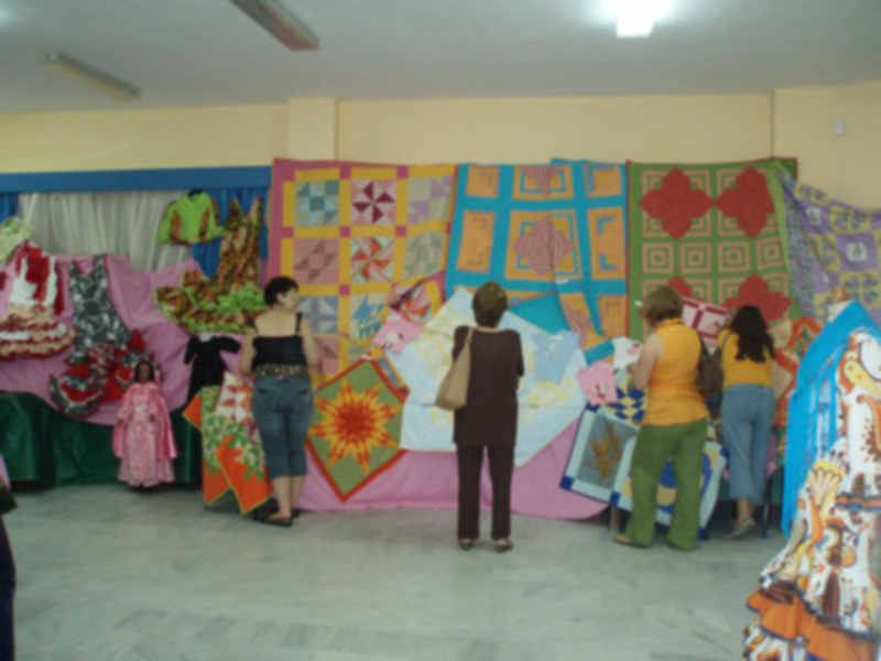Fotografía de una exposición de mantas de colores brillantes y diseños variados, con personas observando y discutiendo sobre las piezas.