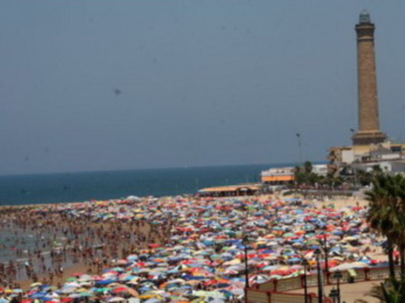 Playa de Chipiona con multitud de bañistas bajo sombrillas coloridas y el faro de Chipiona al fondo.
