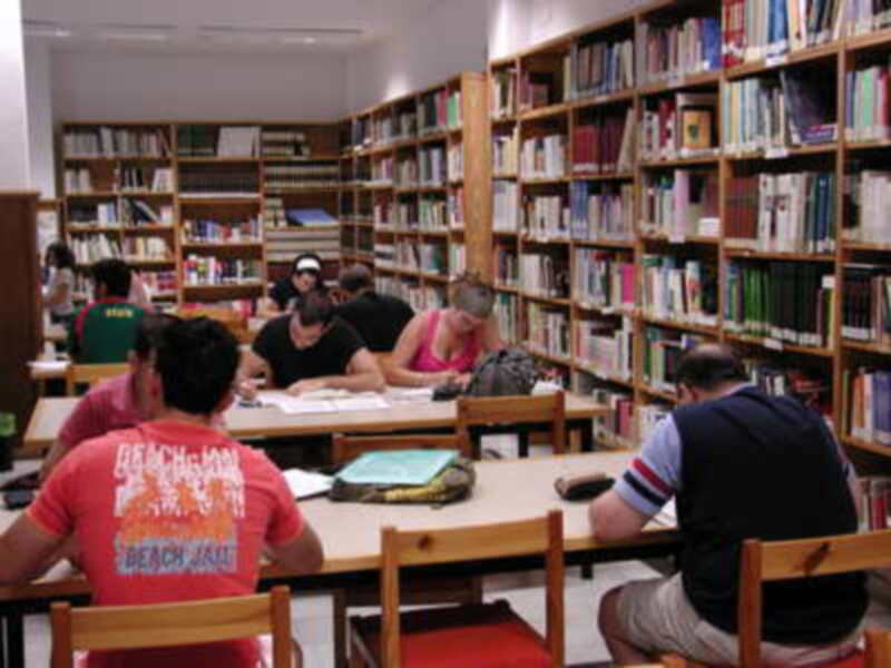 Librería con estanterías llenas de libros. Personas sentadas en mesas, algunos leyendo y otros conversando. Ambiente tranquilo y educativo.