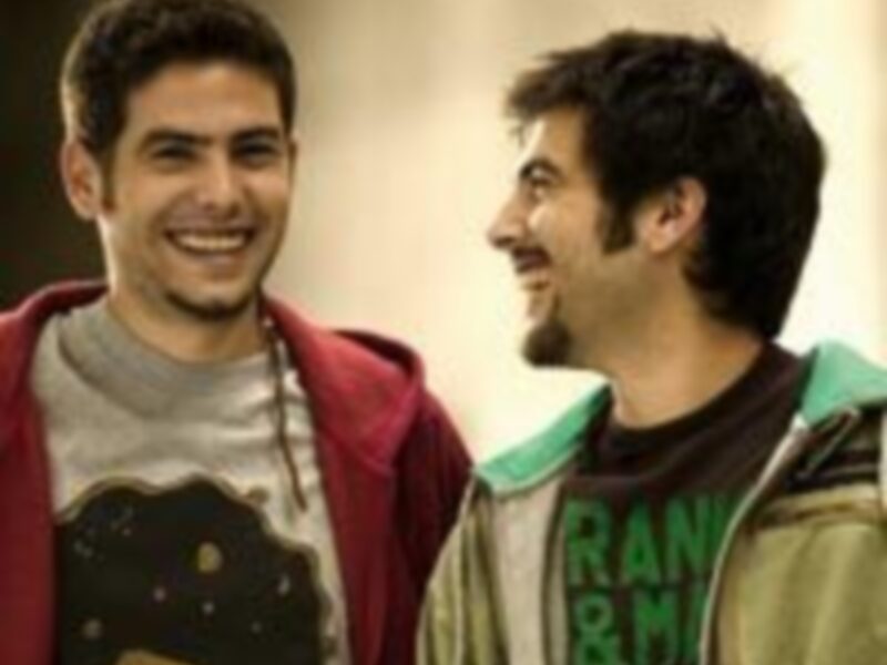 Dos jóvenes sonrientes, uno con una camiseta de dibujos animados y el otro con una camiseta verde con letras negras. Ambos parecen estar en un ambiente casual y relajado, posiblemente una fiesta o evento informal.