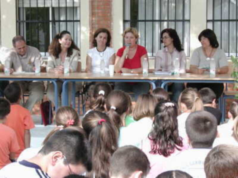 Una escena en un aula con niños sentados frente a una mesa donde se encuentran cuatro mujeres hablando. La imagen muestra un ambiente educativo con estudiantes atentos a las palabras de los adultos.