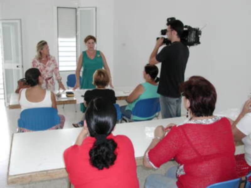 Una mujer en verde se dirige a un grupo de personas sentadas, mientras otro individuo filma la escena con una cámara. La imagen muestra un ambiente de enseñanza o conferencia, con asistentes atentos a la charla.
