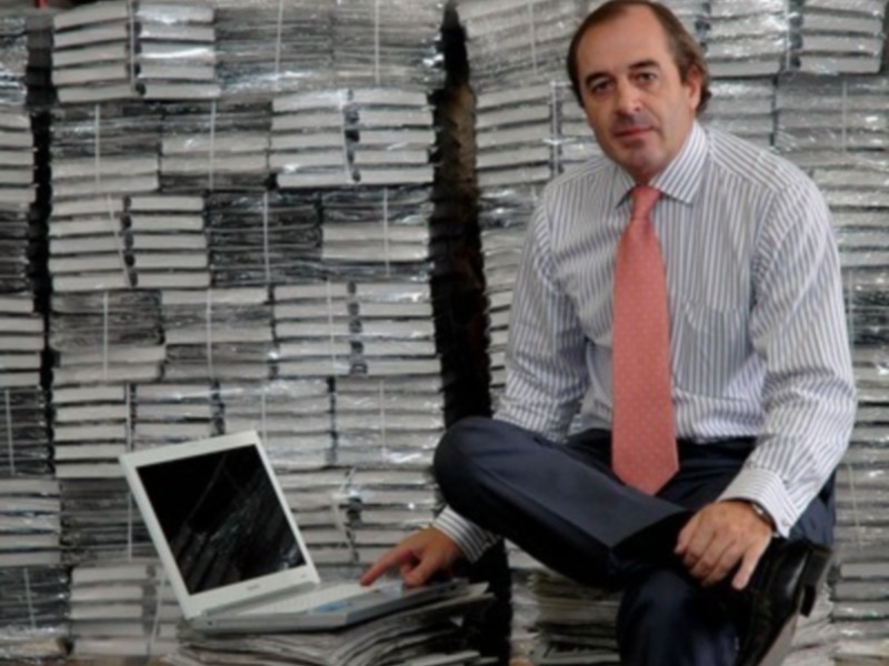Un hombre con corbata rosa y camisa a rayas se sienta en una pila de libros, con un portátil abierto a su lado.