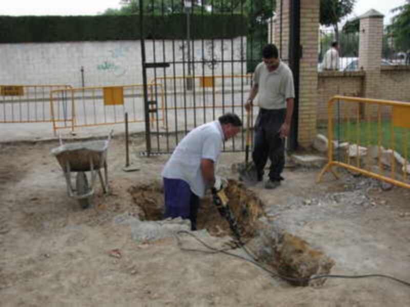 Dos hombres trabajando en la construcción de un muro, con una pala y un martillo. Fondo: valla metálica, cerca de edificios.