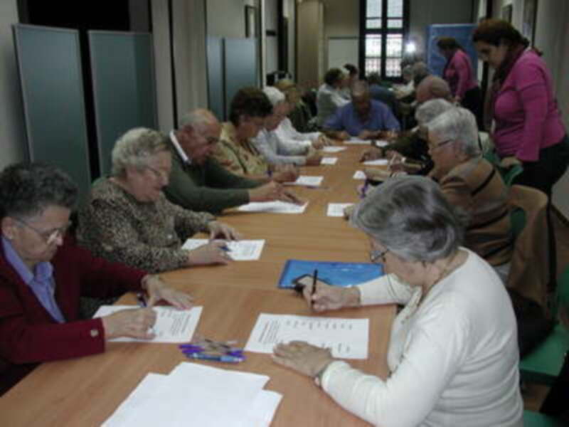 Una reunión de adultos mayores en un salón con mesas y sillas, trabajando en documentos.