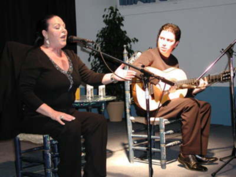 Dos personas se presentan en un escenario, una cantando y la otra tocando guitarra. La escena es informal con sillas de madera y un microtelón en el fondo.