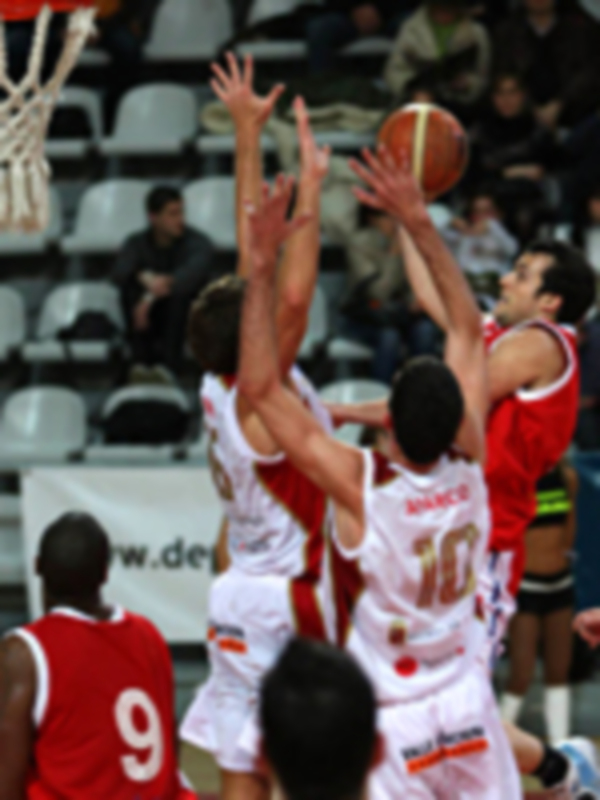 Jugadores en acción durante un partido de baloncesto, con un jugador en rojo intentando bloquear el lanzamiento del jugador en blanco.