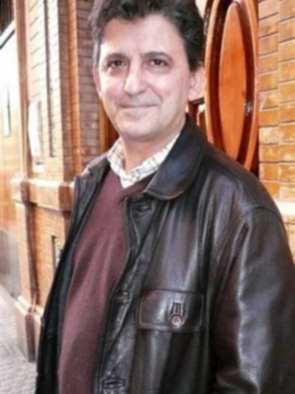 Hombre con chaqueta de cuero y suéter marrón, posando al aire libre frente a una pared de ladrillo.