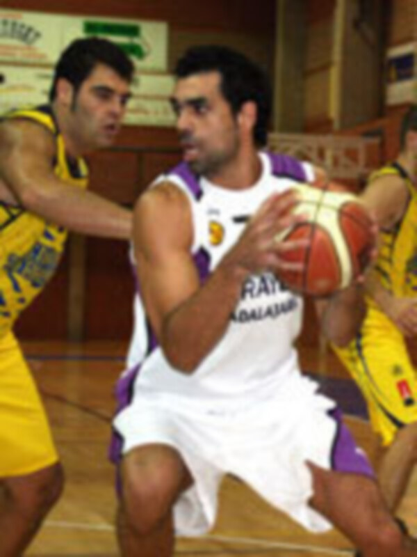 Un jugador de baloncesto en acción durante un partido, con el equipo rival al fondo.