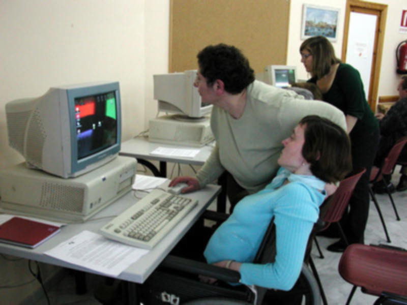 Una persona está sentada en una silla de ruedas, trabajando en un ordenador mientras otra persona observa y ayuda. Ambos están en una sala con varios monitores y computadoras.