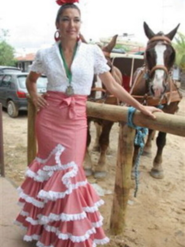 Una mujer con un vestido de flamenca rosa y blanco, posando junto a un caballo en una arena de toros.