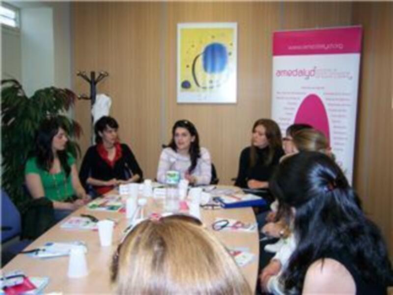 Una reunión de trabajo en un entorno profesional con personas sentadas en una mesa y un cartel de la empresa en el fondo.
