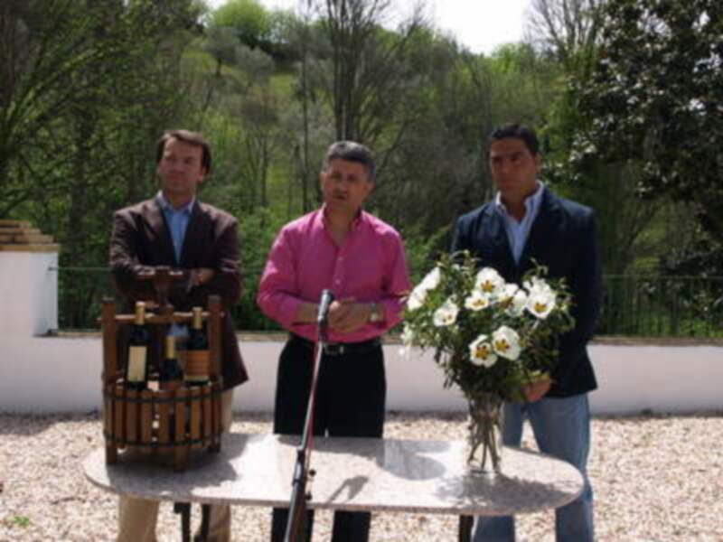 Tres hombres en una presentación al aire libre con botellas de vino y flores en un jardín verde.