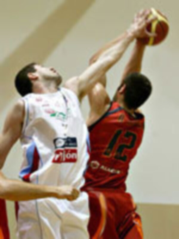 Dos jugadores de baloncesto en acción, uno con el número 12 y otro con el número 3. El jugador con el número 12 lanza la pelota mientras el otro intenta bloquear. Ambos están en un estadio de baloncesto con una pista clara y el fondo es de color claro.