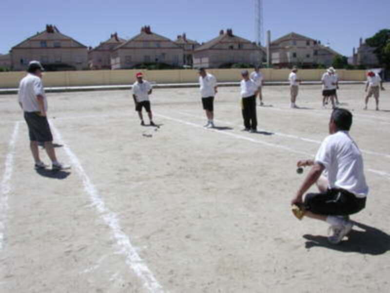Jugadores de bocce en una cancha al aire libre, con edificios en el fondo y un día soleado.
