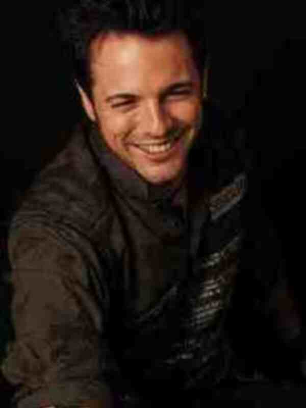 Un hombre sonríe ampliamente, mostrando sus dientes. Lleva una chaqueta negra y un cinturón, con el cabello oscuro. La imagen es de perfil, enfocándose en su expresión facial y la parte superior de su cuerpo.