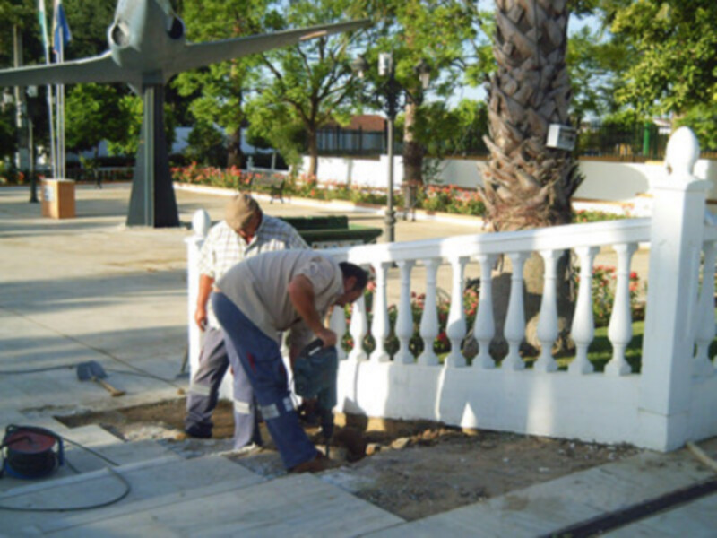 Trabajadores reparando una barandilla en un parque con árboles y flores al fondo.