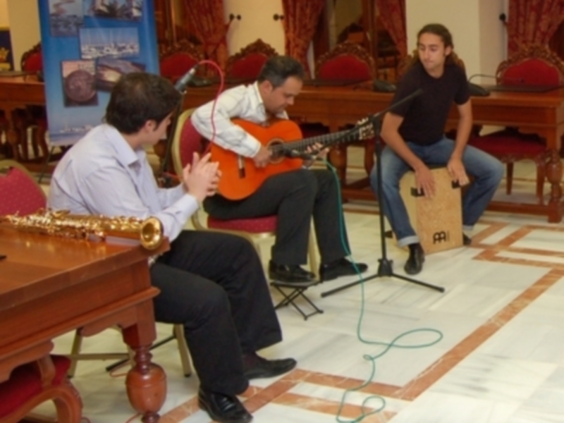 Tres músicos en un ambiente formal, tocando guitarra, saxofón y cajón, con una mesa de madera en el fondo.