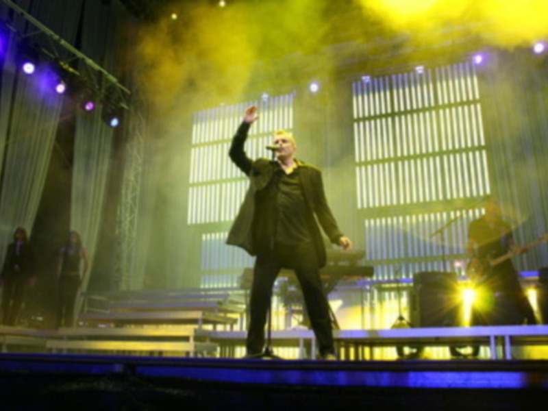 Un hombre en un traje negro se encuentra en el escenario, levantando la mano. La iluminación es intensa con destellos de luz y un fondo que parece ser una ventana con persianas.