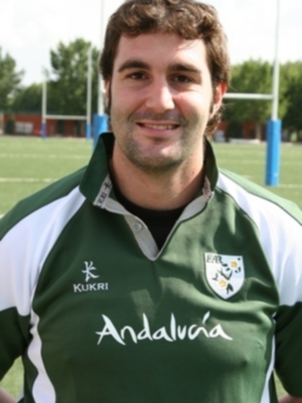 Un hombre con una camiseta verde y blanca de Andalucía, posando en un campo de fútbol con porterías en el fondo.