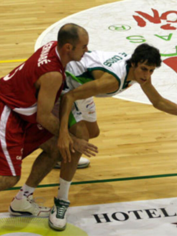 Dos jugadores de baloncesto en acción durante un partido, con un fondo que incluye un logo de hotel y la palabra 'HOTEL' en el suelo del estadio.