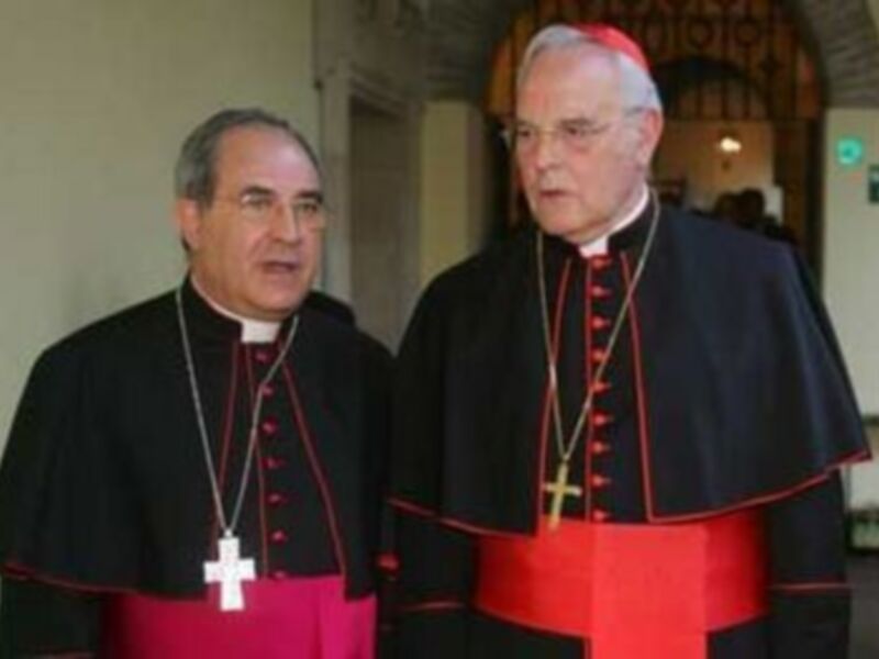Dos sacerdotes en trajes sacerdotales rojos y negros, conversando en un interior bien iluminado.