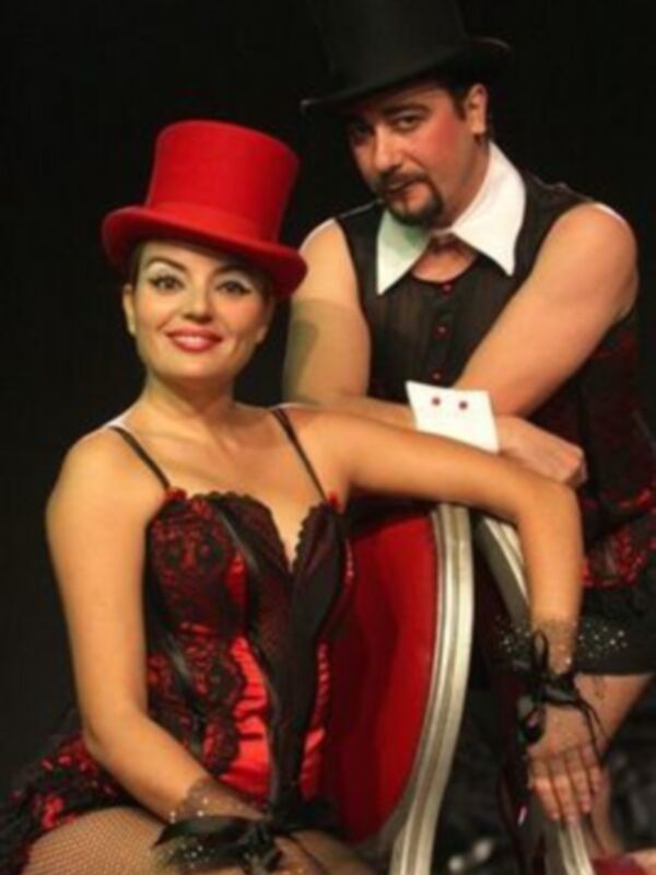 Actores en trajes de circo, una mujer con sombrero rojo y un hombre con traje negro, posando con confianza en un escenario.