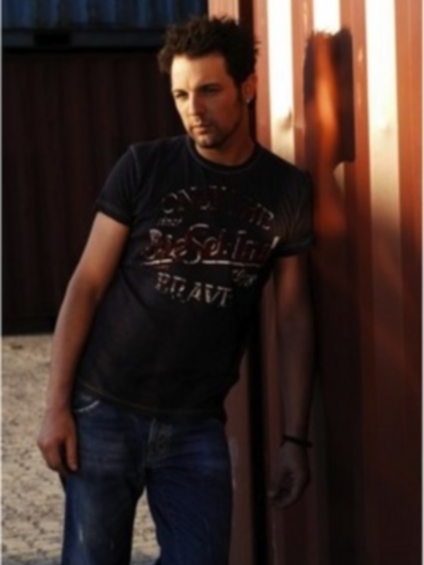 Un hombre joven con cabello oscuro y barba, vestido con una camiseta negra de la banda Rolling Stones y jeans azules, posa contra un muro de hormigón. La luz del sol ilumina su rostro y el fondo está sombreado por un edificio.