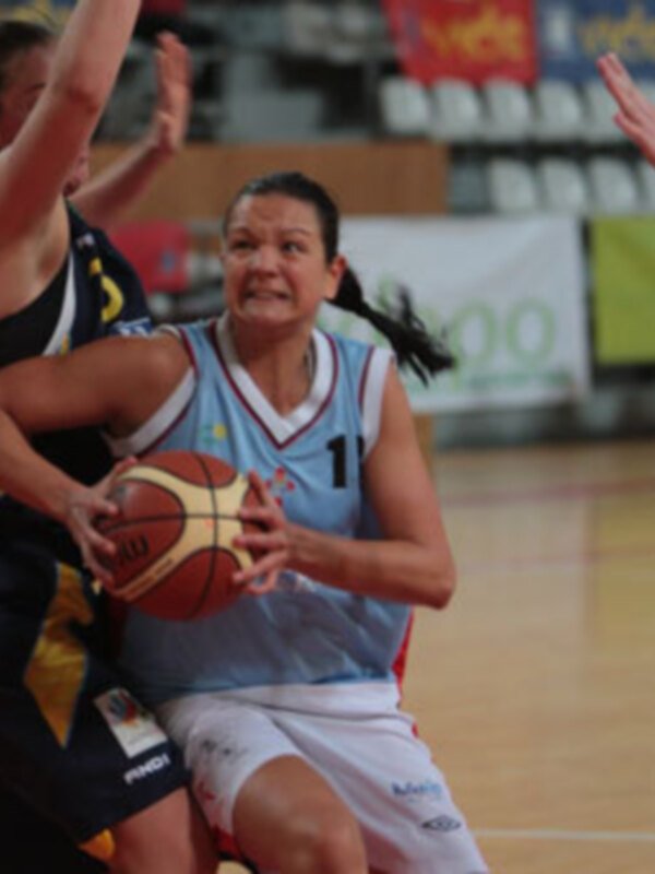 Jugadora de baloncesto en acción durante un partido, con una postura de defensa y una bola en la mano. El fondo muestra una cancha de baloncesto y espectadores en los gradas.