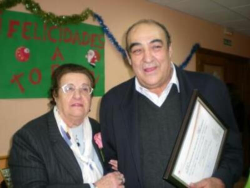 Dos personas posan juntas, una con un diploma y la otra con un ramo de flores. La imagen se encuentra en un ambiente festivo, con una pancarta que dice "Felicidades a tod".